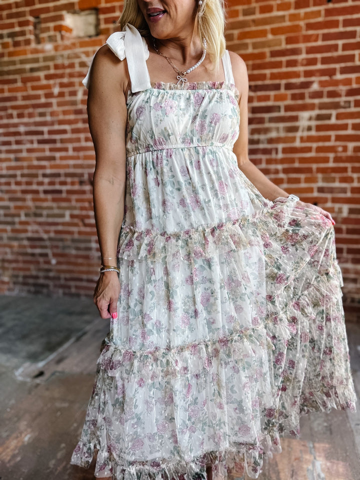 A woman wearing a floral chiffon dress with tie straps and a tiered silhouette, standing against a brick wall.
