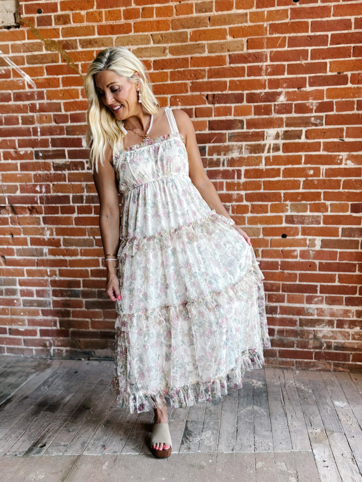 A woman wearing a floral chiffon dress with tie straps and a tiered silhouette, standing against a brick wall.