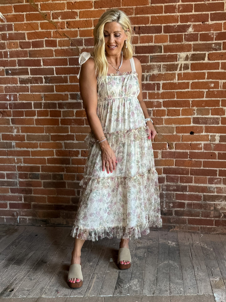 A woman wearing a floral chiffon dress with tie straps and a tiered silhouette, standing against a brick wall.