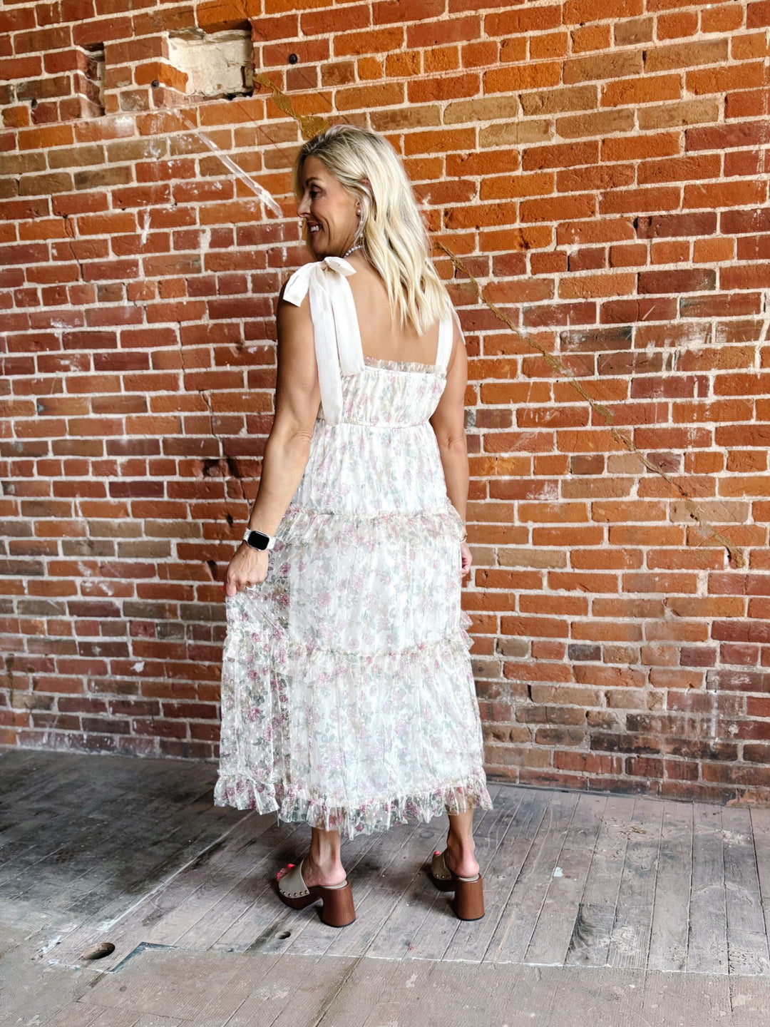 A woman wearing a floral chiffon dress with tie straps and a tiered silhouette, standing against a brick wall.