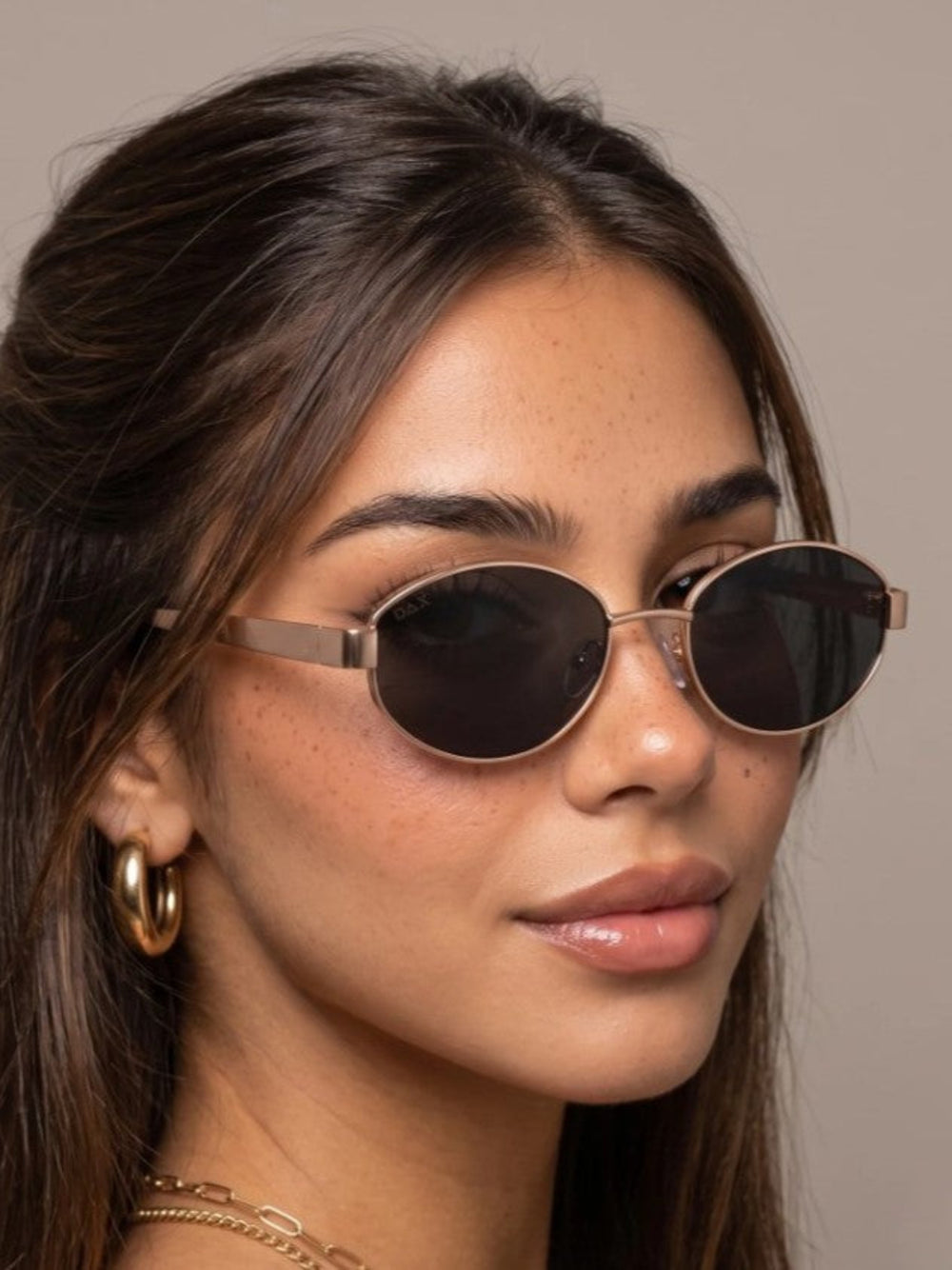 A close-up of a woman wearing oval sunglasses with dark lenses and thin rose gold metal frames, paired with gold hoop earrings and a delicate gold chain necklace, set against a solid taupe background.