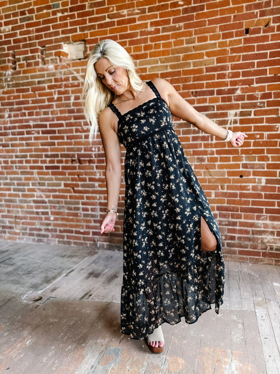 A woman wearing a sleeveless floral midi dress with lace trim, standing against a brick wall.