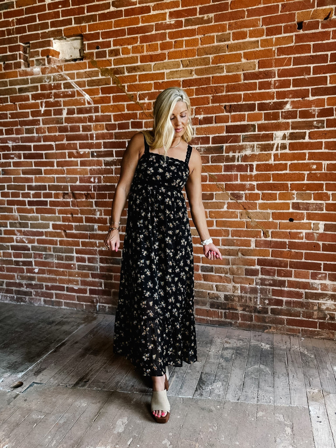 A woman wearing a sleeveless floral midi dress with lace trim, standing against a brick wall.