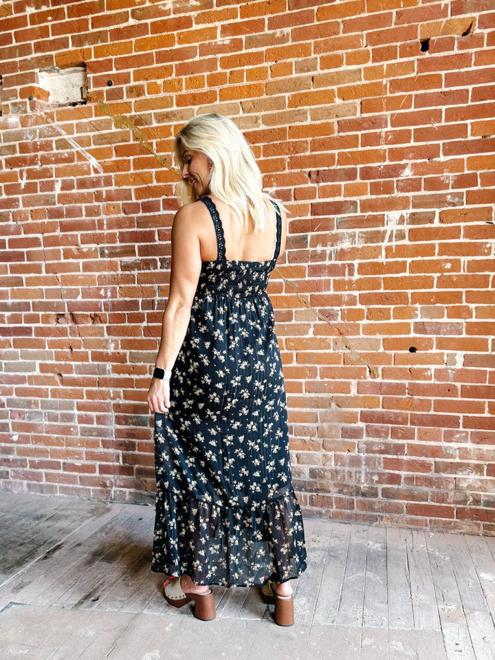 A woman wearing a sleeveless floral midi dress with lace trim, standing against a brick wall.