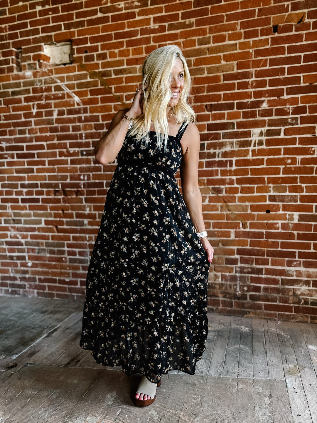 A woman wearing a sleeveless floral midi dress with lace trim, standing against a brick wall.