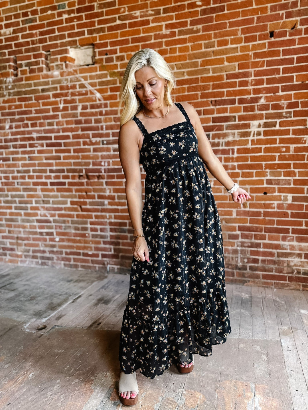 A woman wearing a sleeveless floral midi dress with lace trim, standing against a brick wall.
