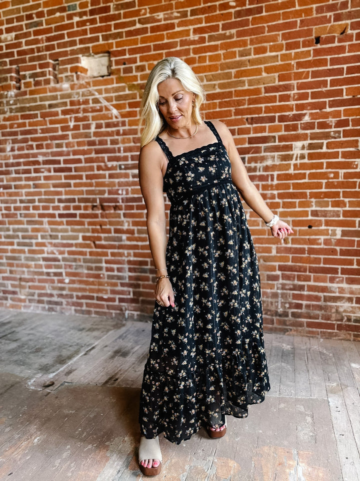 A woman wearing a sleeveless floral midi dress with lace trim, standing against a brick wall.