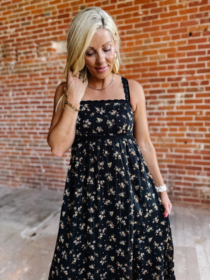 A woman wearing a sleeveless floral midi dress with lace trim, standing against a brick wall.