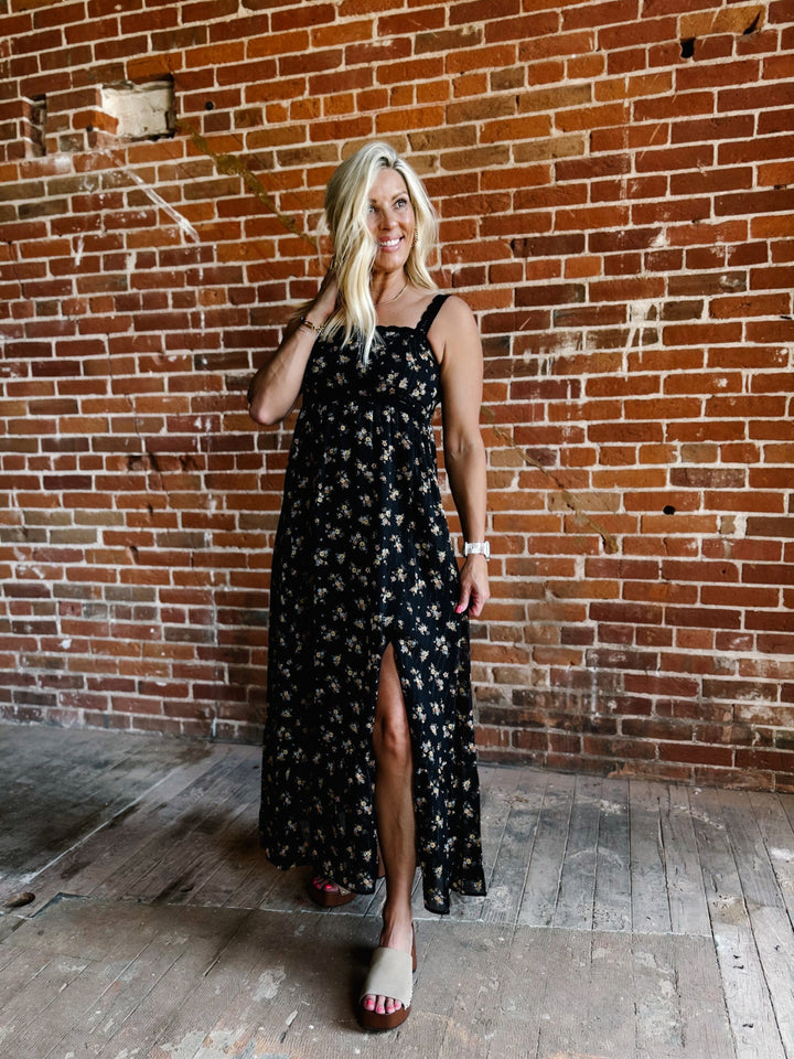 A woman wearing a sleeveless floral midi dress with lace trim, standing against a brick wall.