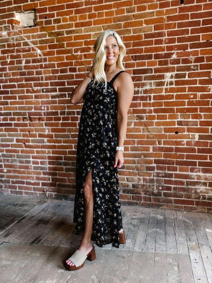 A woman wearing a sleeveless floral midi dress with lace trim, standing against a brick wall.