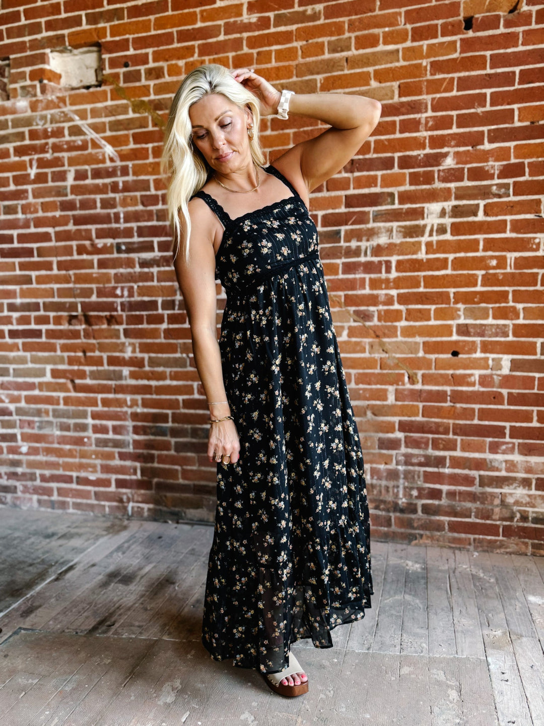 A woman wearing a sleeveless floral midi dress with lace trim, standing against a brick wall.