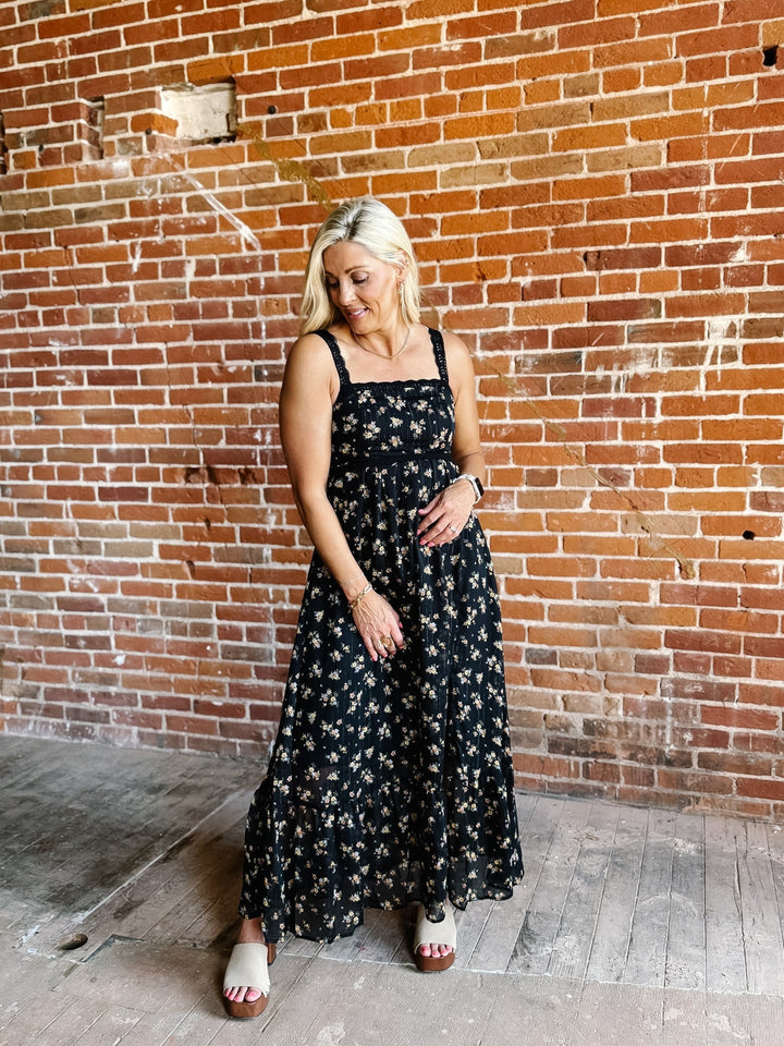 A woman wearing a sleeveless floral midi dress with lace trim, standing against a brick wall.