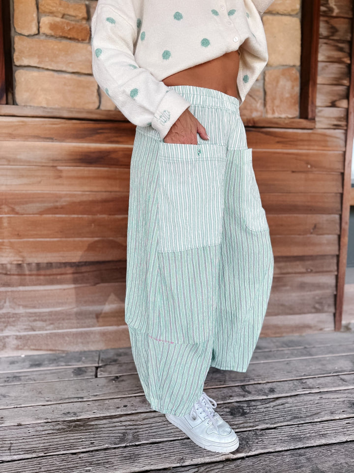 A person wearing light mint green and white vertically striped wide-leg pants, paired with a cream cardigan and white sneakers, standing on a wooden deck in front of a rustic wood and stone wall.
