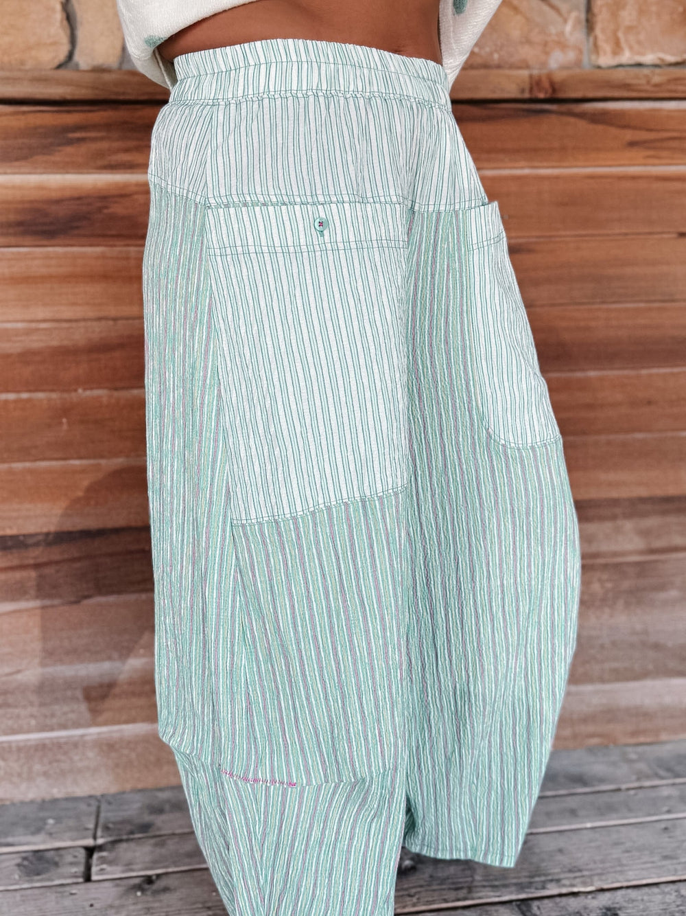 A close-up of a person wearing high-waisted, wide-leg striped pants in mint green and white, standing against a rustic wooden plank wall.