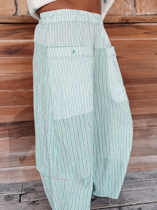A close-up of a person wearing high-waisted, wide-leg striped pants in mint green and white, standing against a rustic wooden plank wall.