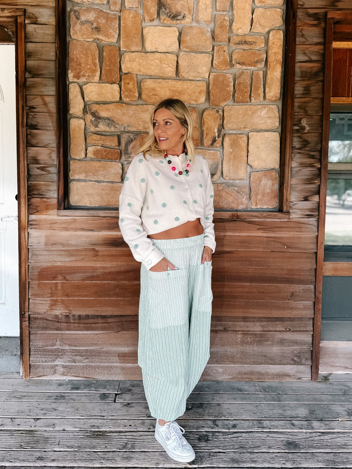 A woman stands on a rustic wooden porch in front of a stone wall, wearing a cream cropped sweater with mint green dots, light green pinstriped wide-leg pants, and white sneakers.
