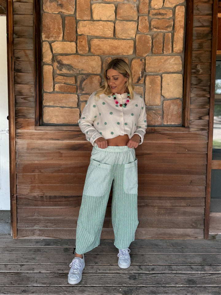 A woman stands on a wooden deck in front of a rustic stone wall, wearing a cream long-sleeve crop top with green polka dots, mint green and white vertically striped wide-leg pants, and white sneakers.
