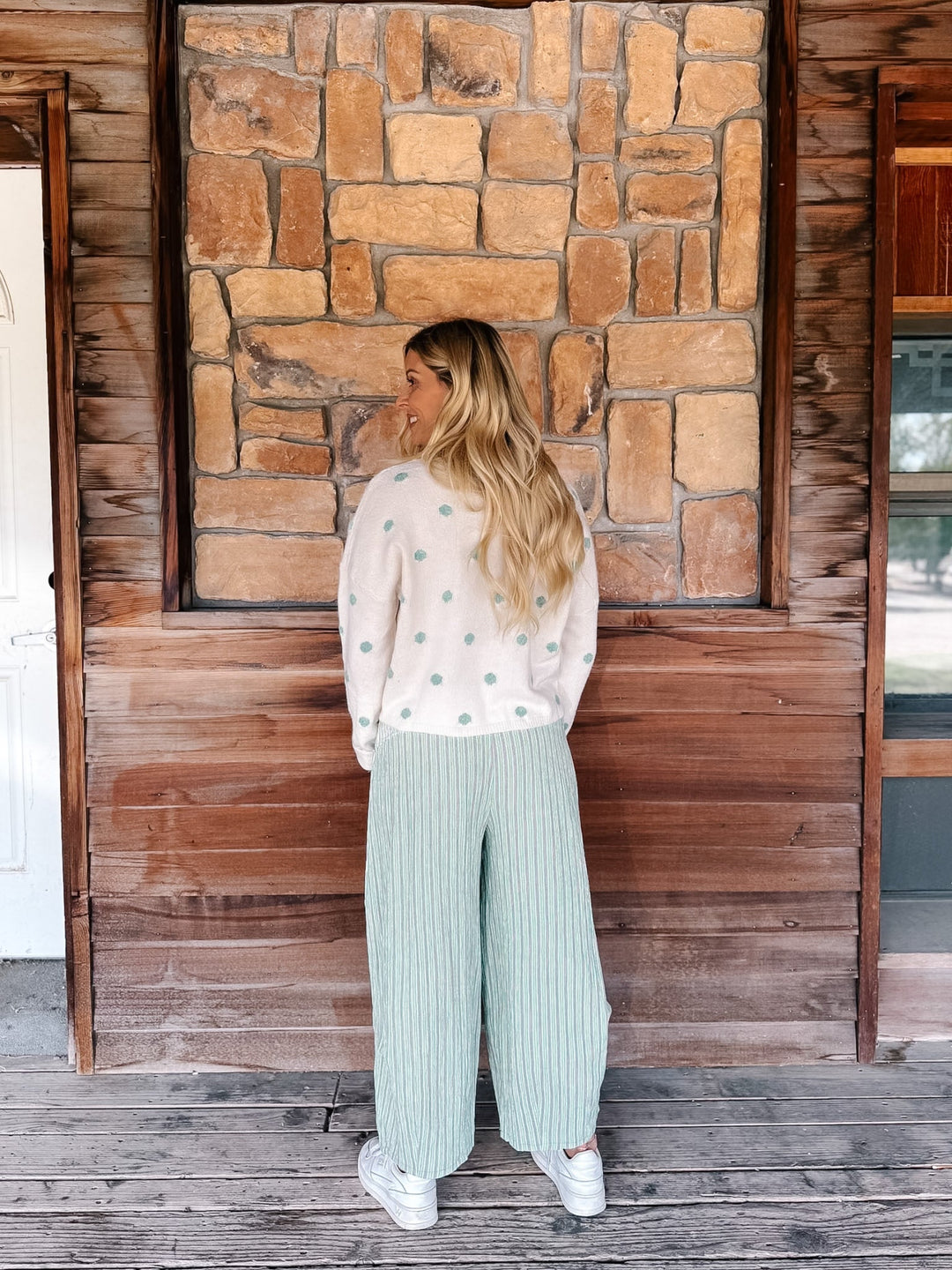 A woman stands on a rustic wooden porch facing a stone wall, wearing a cream knit sweater with mint green polka dots and matching wide-leg pleated pants.