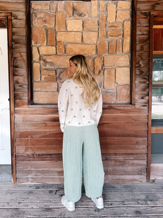 A woman stands on a rustic wooden porch facing a stone wall, wearing a cream knit sweater with mint green polka dots and matching wide-leg pleated pants.