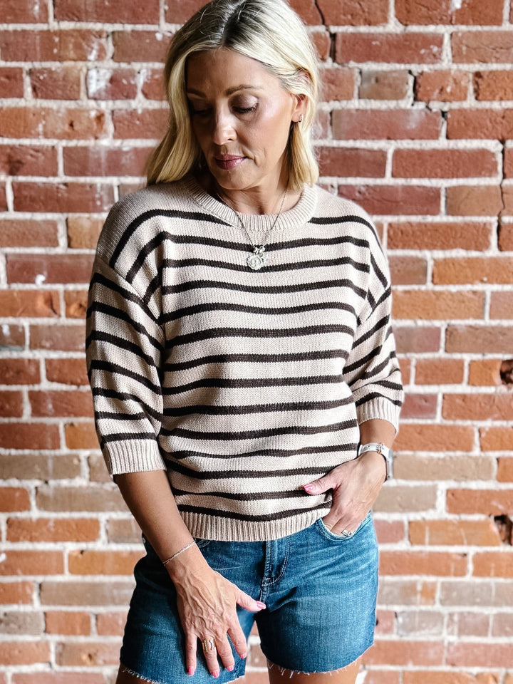 A woman wearing a short sleeve sweater top with mocha stripes and a crew neckline, paired with denim shorts.