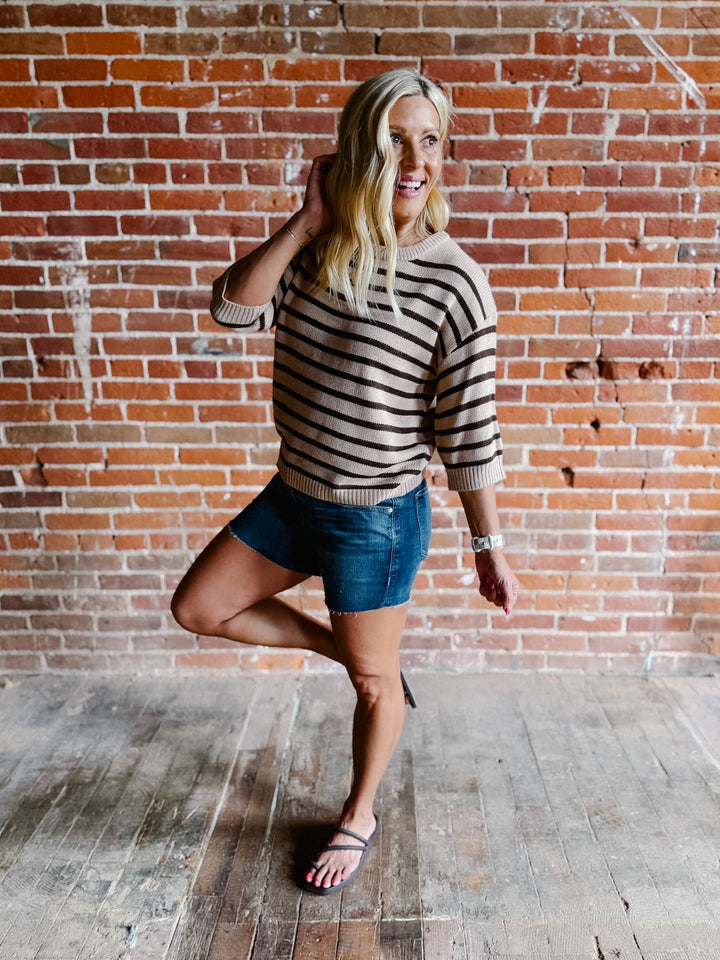 A woman wearing a short sleeve sweater top with mocha stripes and a crew neckline, paired with denim shorts.