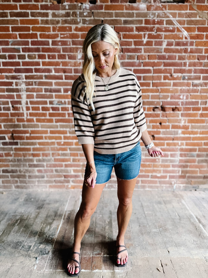 A woman wearing a short sleeve sweater top with mocha stripes and a crew neckline, paired with denim shorts.