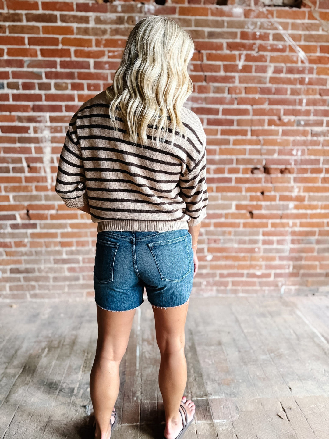 A woman wearing a short sleeve sweater top with mocha stripes and a crew neckline, paired with denim shorts.
