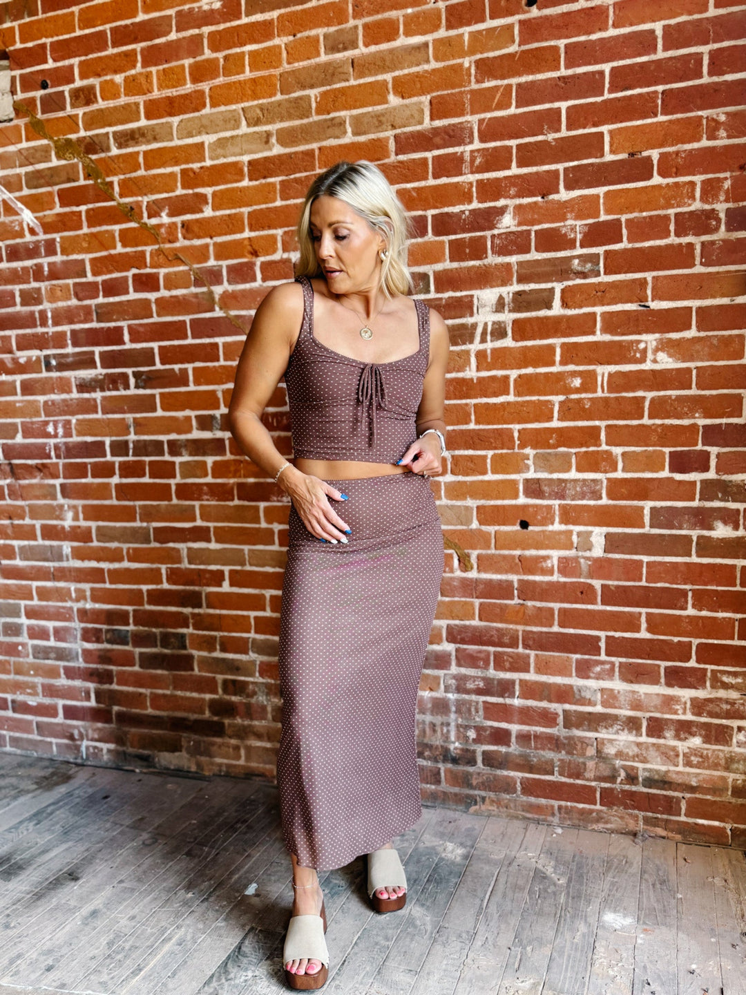 A woman modeling a matching two-piece set consisting of a brown textured crop top with a tie detail and a high-waisted maxi skirt, set against a rustic red brick wall.