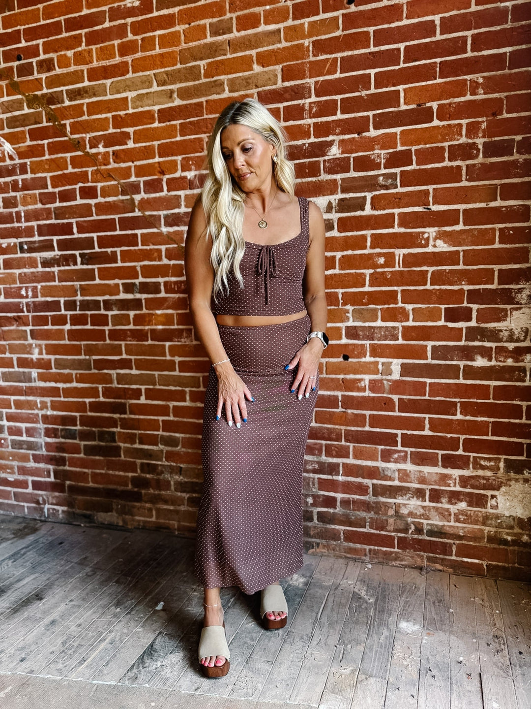 A woman modeling a matching two-piece set consisting of a brown polka dot crop top and a long brown polka dot skirt, standing against a red brick wall.