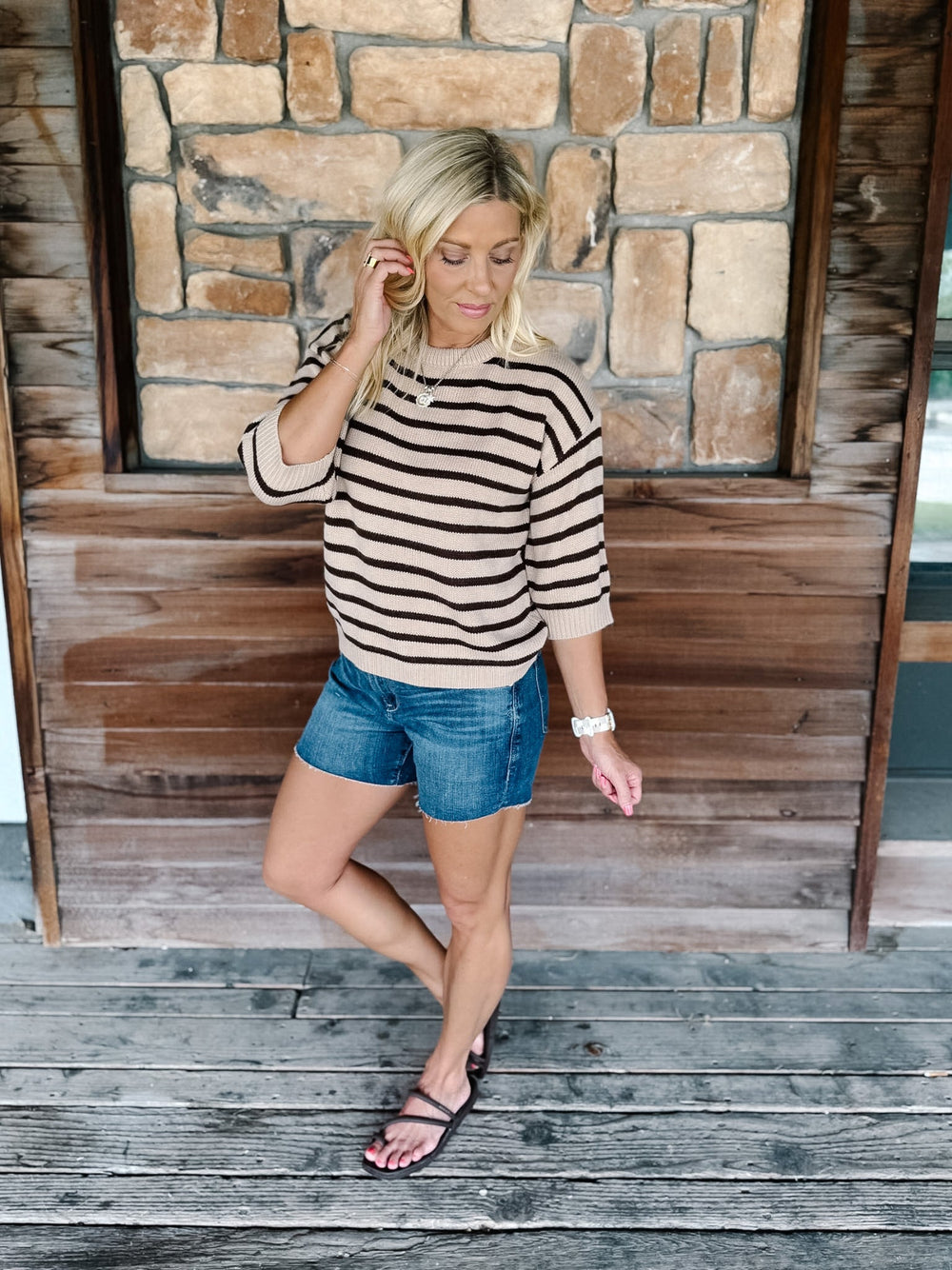 A woman stands on a rustic wooden porch in front of a stone wall, wearing a beige and black horizontally striped knit sweater, blue denim shorts, and black slide sandals.