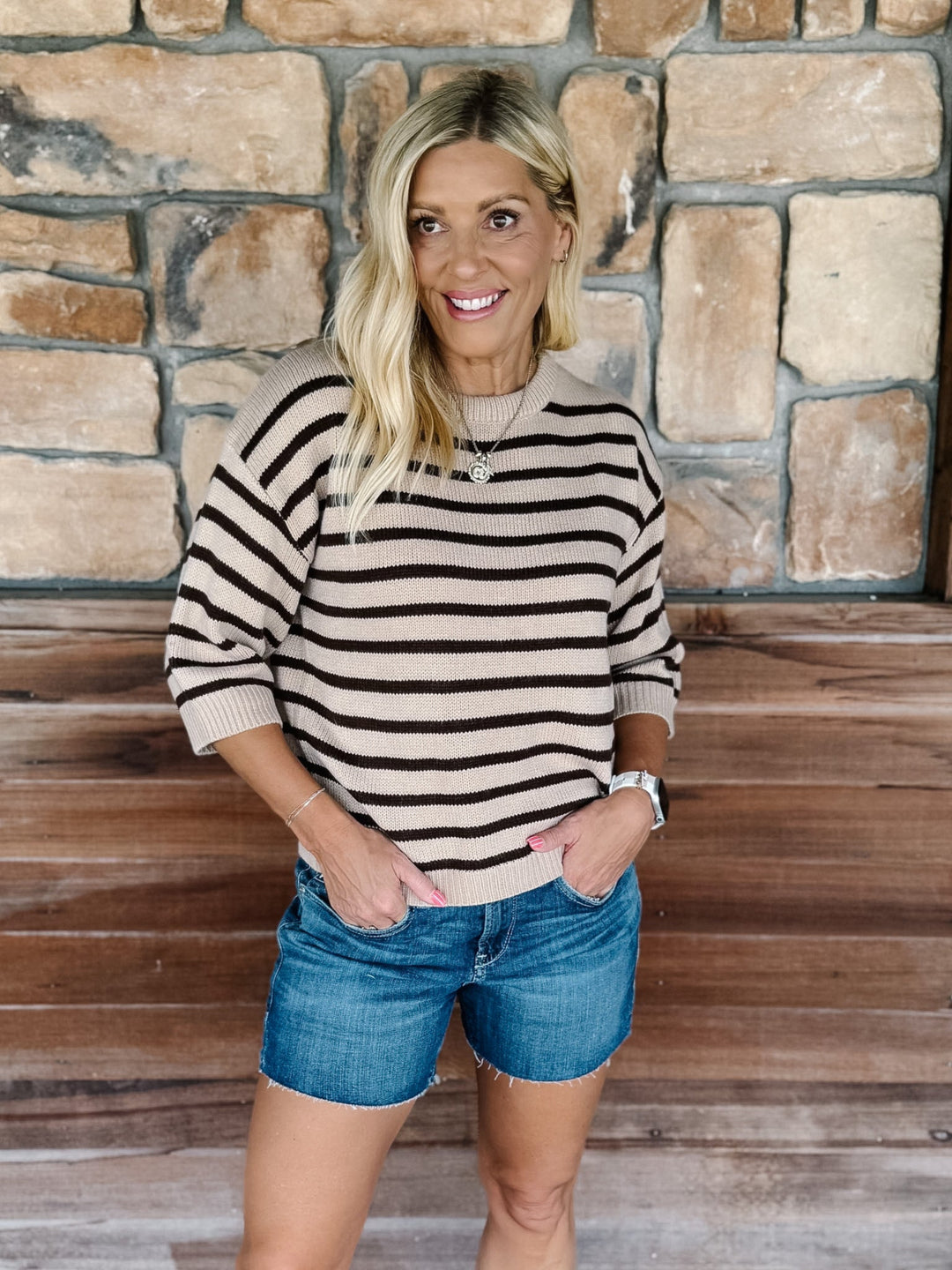 A smiling blonde woman stands in front of a stone wall and wooden steps, wearing a beige knit sweater with dark brown horizontal stripes and blue denim shorts.
