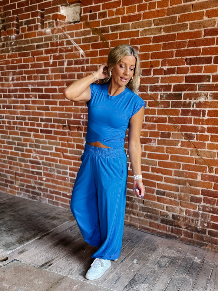 A woman modeling a bright blue two-piece activewear set consisting of a short-sleeve crop top and matching wide-leg pants, standing against a red brick wall.