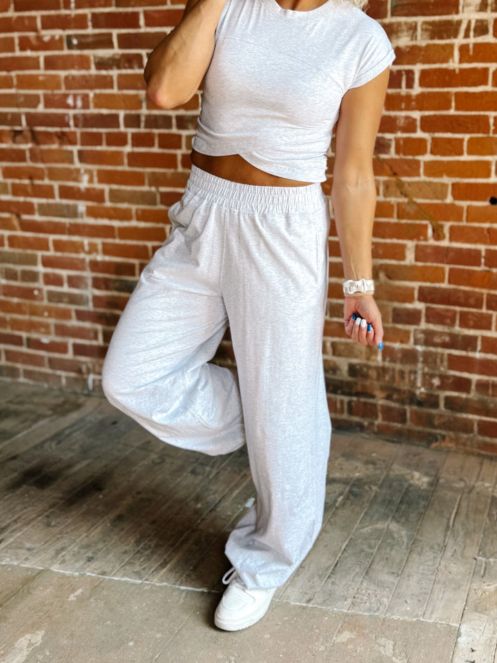 A person wearing a light gray heather crop top and matching wide-leg sweatpants, standing in front of a brick wall and concrete floor.