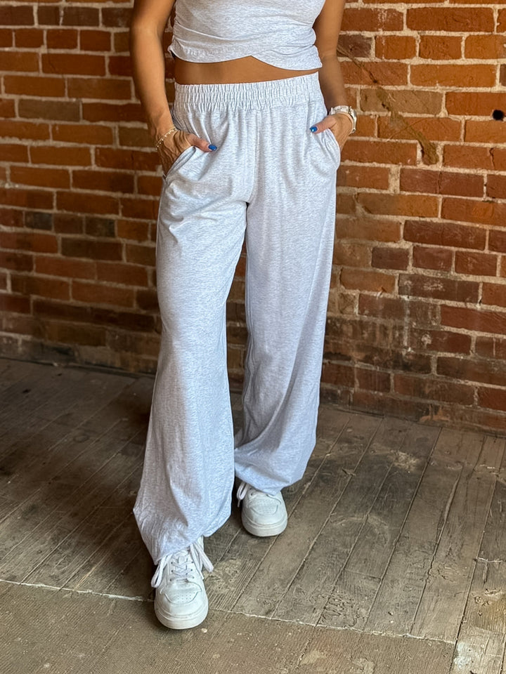 A person wearing light heather gray wide-leg sweatpants with an elastic waistband and side pockets, paired with a matching light gray crop top and white sneakers, standing against a brick wall.