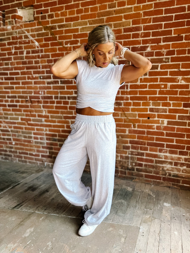 A woman posing in a white crop top and matching white wide-leg sweatpants against a red brick wall background.