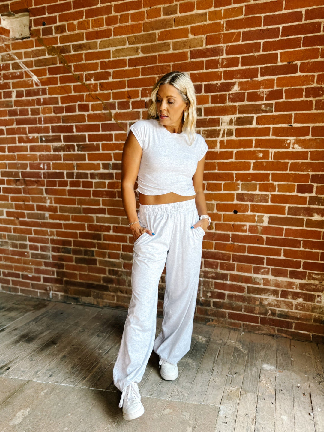 A woman posing in a white short-sleeve crop top and matching white wide-leg sweatpants against a rustic red brick wall.
