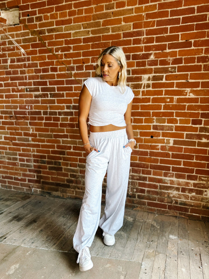 A woman posing in a white short-sleeve crop top and matching white wide-leg sweatpants against a rustic red brick wall.
