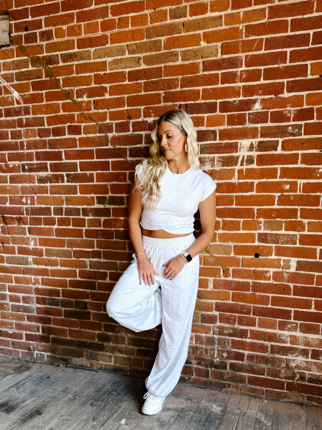 A woman posing in a white crop top and matching white sweatpants against a red-brown brick wall.