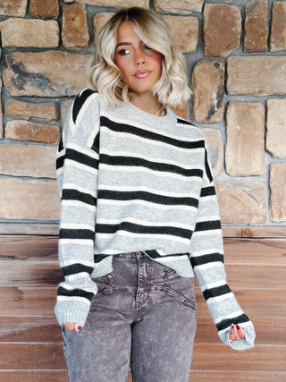 A woman wearing a light gray and Brown horizontally striped knit sweater, paired with brown jeans, standing against a rustic stone wall.