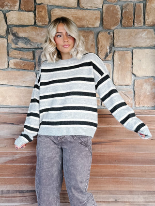 A woman wearing a light gray and Brown horizontally striped knit sweater, paired with brown jeans, standing against a rustic stone wall.