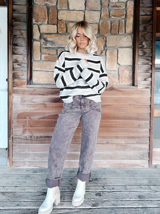 A woman wearing a light gray and Brown horizontally striped knit sweater, paired with brown jeans, standing against a rustic stone wall.