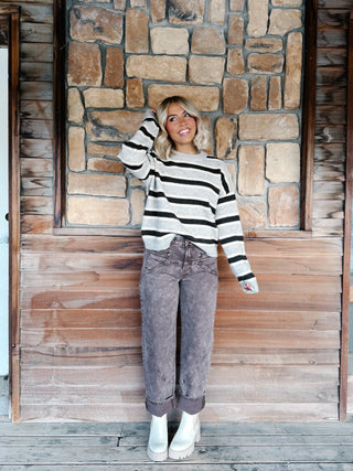 A woman wearing a light gray and Brown horizontally striped knit sweater, paired with brown jeans, standing against a rustic stone wall.