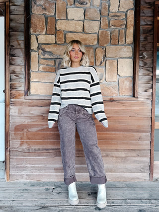 A woman wearing a light gray and Brown horizontally striped knit sweater, paired with brown jeans, standing against a rustic stone wall.