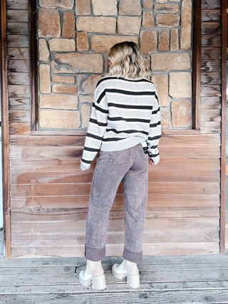 A woman wearing a light gray and Brown horizontally striped knit sweater, paired with brown jeans, standing against a rustic stone wall.