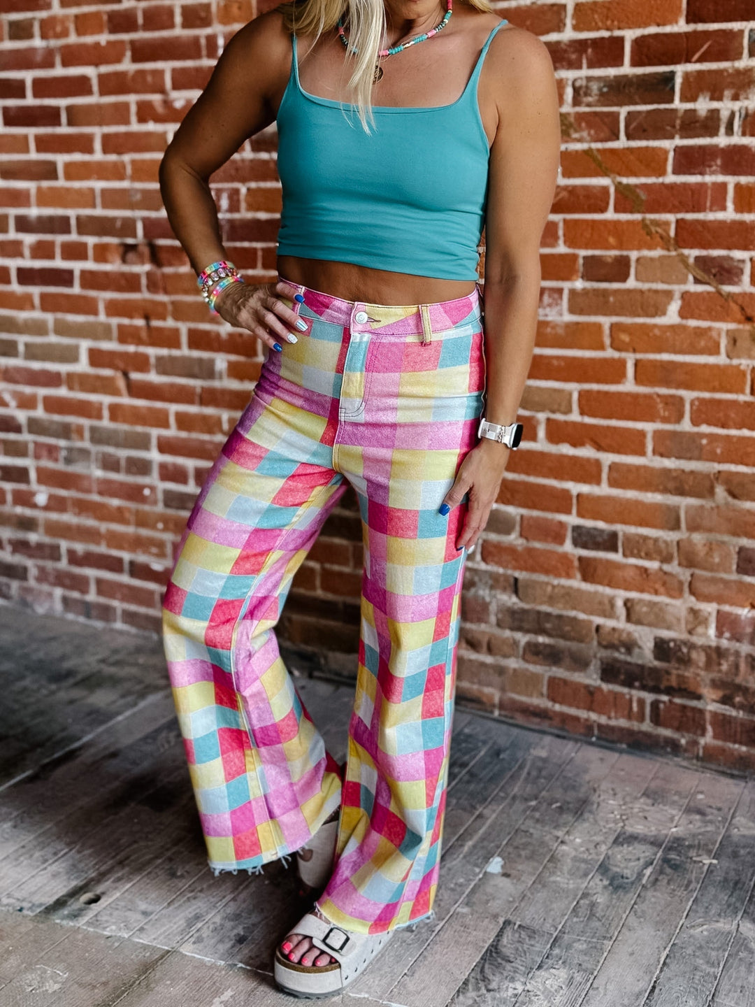 A woman models a two-piece outfit featuring a teal crop top and high-waisted, wide-leg plaid pants in pink, yellow, and blue, standing against a rustic brick wall.