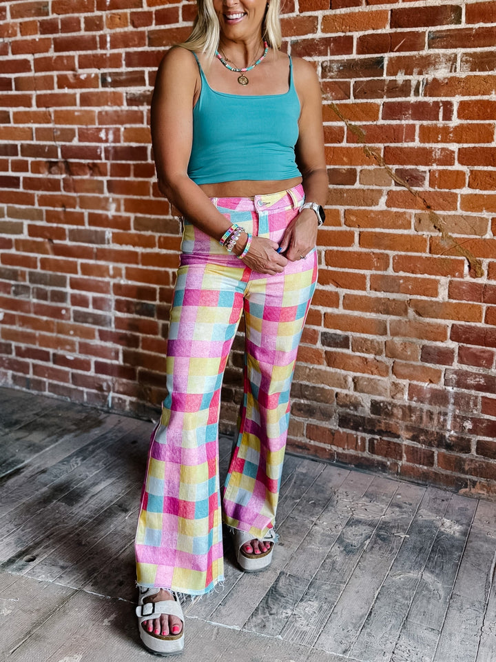 A woman wearing a teal crop top and pastel multicolor plaid flare pants, standing against a red brick wall.