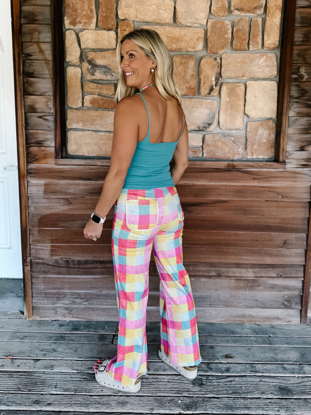 A woman stands on a rustic wooden porch in front of a stone wall, wearing pastel plaid wide-leg pants with a teal tank top and white sneakers.