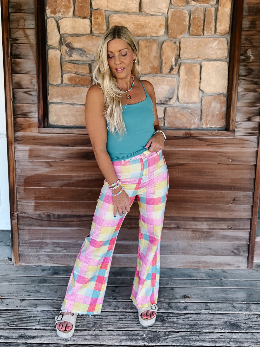 A woman stands on a wooden porch in front of a stone wall, wearing pastel plaid wide-leg pants with pink, yellow, and blue tones, a teal tank top, and white slide sandals.