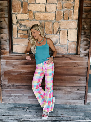 A woman posing in front of a rustic stone and wood wall, wearing a teal tank top and pastel pink, yellow, and blue plaid wide-leg pants.