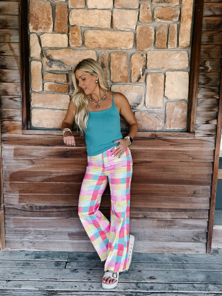 A woman posing in front of a rustic stone and wood wall, wearing a teal tank top and pastel pink, yellow, and blue plaid wide-leg pants.